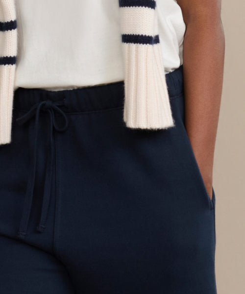 A person wears Bay Sweatpant navy blue drawstring pants, a white top, and a white scarf with navy stripes, with one hand in their pocket.