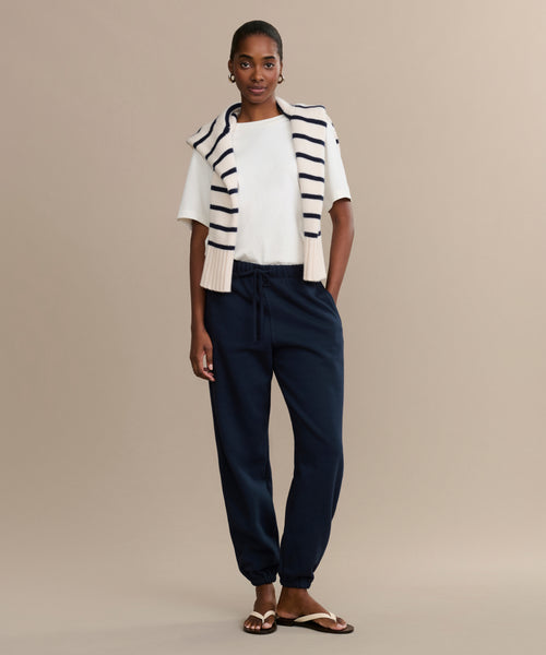 Against a beige background, a woman wears a white T-shirt, navy Bay Sweatpant joggers in brushed fleece, sandals, and has a white sweater with navy stripes draped over her shoulders, with her hands in her pockets.