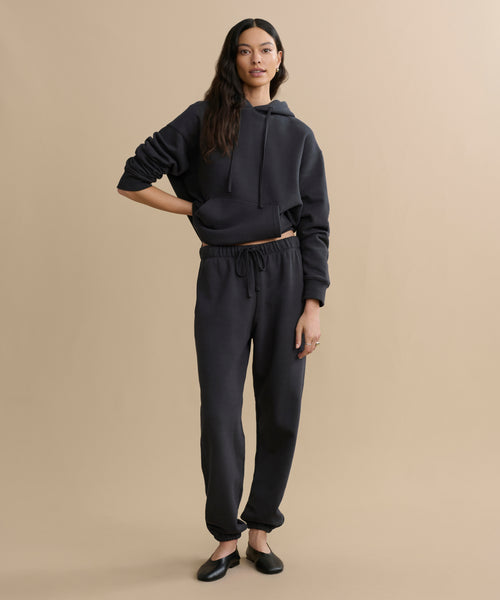 A woman with long dark hair poses casually against a beige background, wearing a black hoodie and the Bay Sweatpant in athletic brushed fleece, paired with black flats.