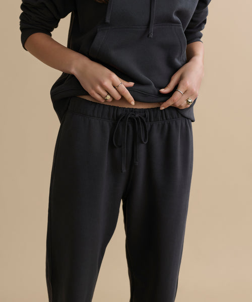 A person in a black hoodie and Bay Sweatpant brushed fleece athletic sweats stands against a beige background, adjusting the waistband. Their face is not visible.