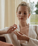 A blonde-haired woman in a cream cardigan sits by a window indoors, calmly examining a small jar from the Best-Selling Balm Duo.