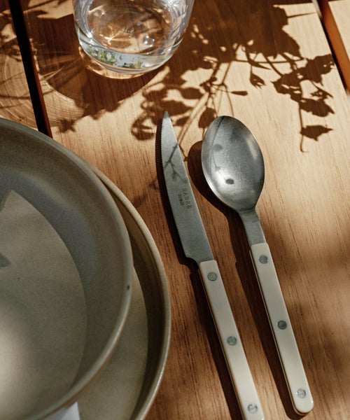 A Bistro Vintage Finish Cutlery Set with white handles rests on a wooden table next to stacked ceramic plates and a glass of water. Sunlight and leaf shadows highlight the charm of this elegant cutlery.