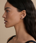 A woman with long dark hair is shown in profile against a beige background, wearing a black top, a Jenni Kayne Jewelry black stud earring in her earlobe, and the Loren Pearl Ear Cuff on her upper ear.