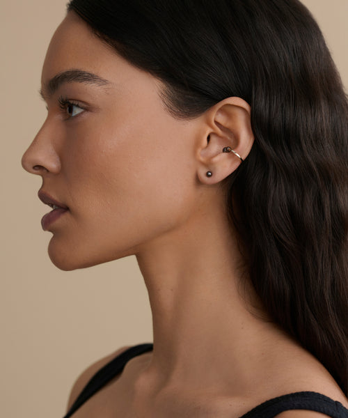 A woman with long dark hair is shown in profile against a beige background, wearing a black top, a Jenni Kayne Jewelry black stud earring in her earlobe, and the Loren Pearl Ear Cuff on her upper ear.