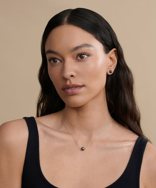 A woman with long dark hair, in a black sleeveless top, small stud earring, and the Philo Pearl Necklace—a delicate black pearl necklace with a round pendant—poses against a plain beige background.