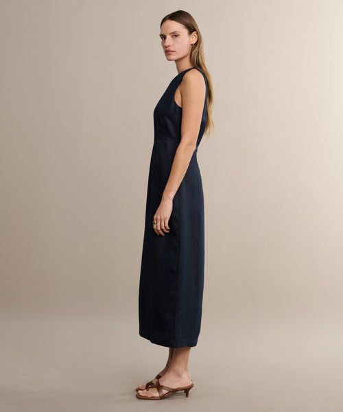 A woman with long brown hair stands in profile, wearing the Blair Dress—sleeveless, ankle-length with a subtle linen texture—and brown heeled sandals against a plain, light beige background.