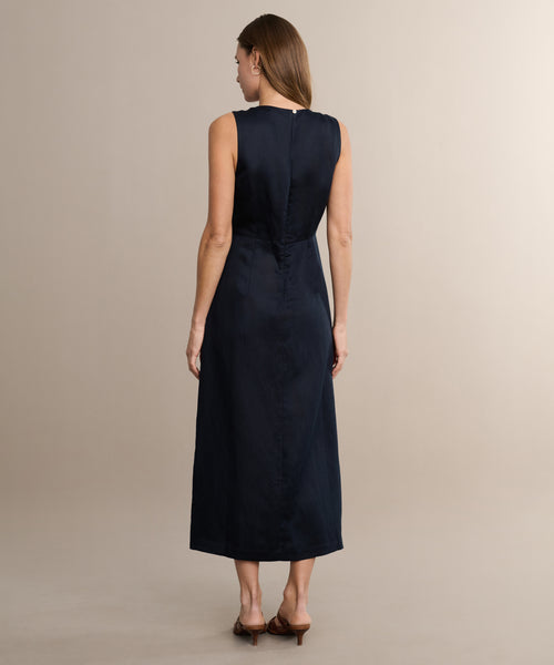 Woman stands with her back to the camera, wearing the sleeveless, ankle-length Blair Dress in dark navy linen and brown heeled sandals against a neutral beige background—an ideal choice to refresh your spring wardrobe.