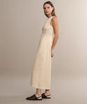 A woman stands in profile against a beige background wearing the Blair Dress, a sleeveless, ankle-length cream linen piece perfect for spring, styled with black woven flats.
