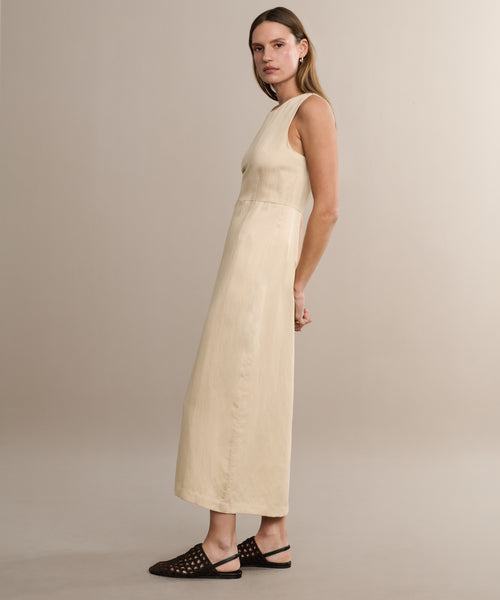 A woman stands in profile against a beige background wearing the Blair Dress, a sleeveless, ankle-length cream linen piece perfect for spring, styled with black woven flats.