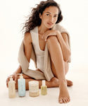 A woman with long wavy hair sits barefoot on the floor in a white outfit and beige sweater, surrounded by Oak Essentials The Body Routine skincare products, including bottles, a jar, and a dropper bottle.