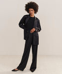 A woman with an afro hairstyle wears the versatile Bond Blazer, a black top, wide-leg black trousers, and black heels, standing on a light carpet against a plain beige background.