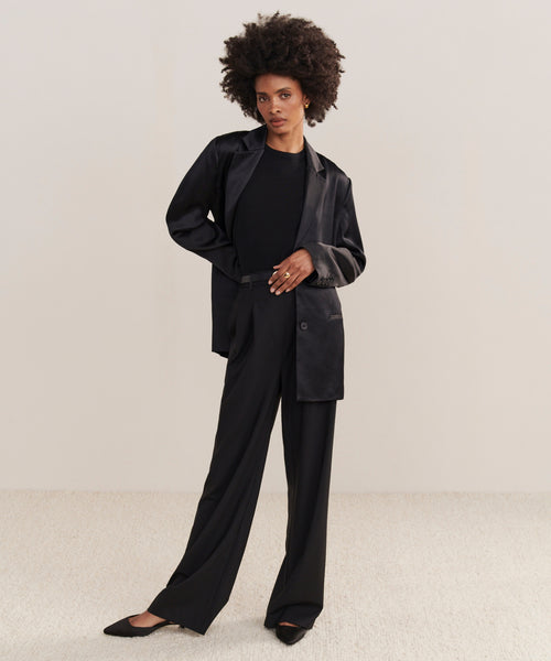A woman with an afro hairstyle wears the versatile Bond Blazer, a black top, wide-leg black trousers, and black heels, standing on a light carpet against a plain beige background.