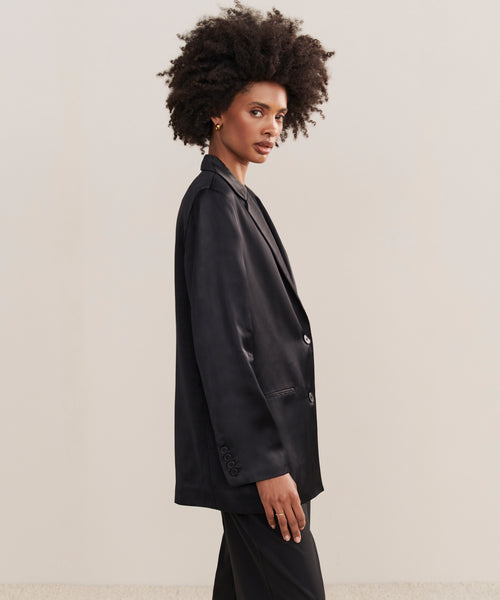 A woman with natural curly hair models the Bond Blazer and matching pants, standing sideways and looking at the camera against a plain, light background.