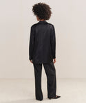 A person with curly hair faces away, wearing the Bond Blazer in loose-fitting black viscose satin with matching pants and black pointed-toe shoes, set against a plain light background.