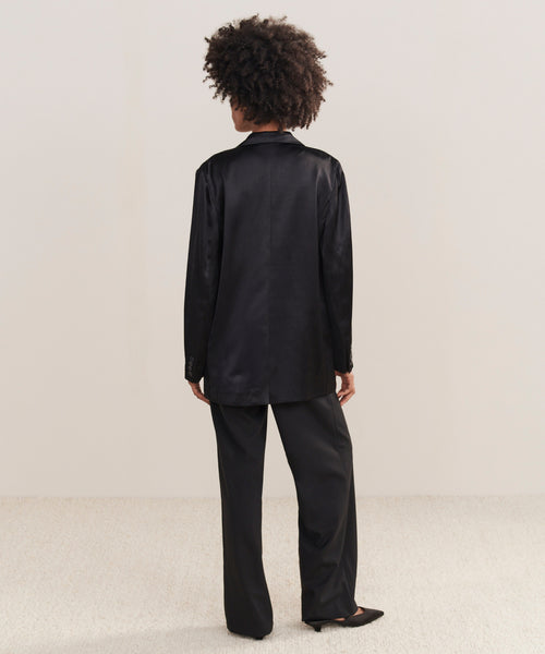 A person with curly hair faces away, wearing the Bond Blazer in loose-fitting black viscose satin with matching pants and black pointed-toe shoes, set against a plain light background.