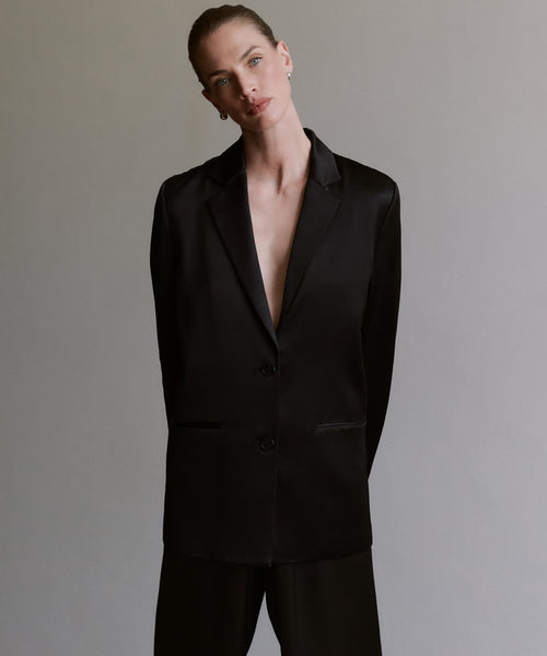 A person with slicked-back hair wears the Bond Blazer with no shirt underneath and matching black pants, standing against a plain gray background with a neutral expression.