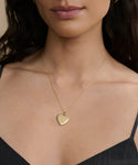 A woman wears the Bond Locket, a vintage-inspired gold heart necklace with a small gemstone, paired with a black top. The image highlights her neckline to showcase the elegant jewelry.