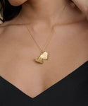 A woman wears the Bond Locket, a vintage-inspired gold heart necklace on a delicate chain, with the locket slightly open against her bare skin and a dark V-neck top.