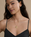 A woman with long dark hair wears a black spaghetti-strap top and the Bond Locket, posing against a beige background. The vintage-inspired Bond Locket adds timeless elegance to her look.