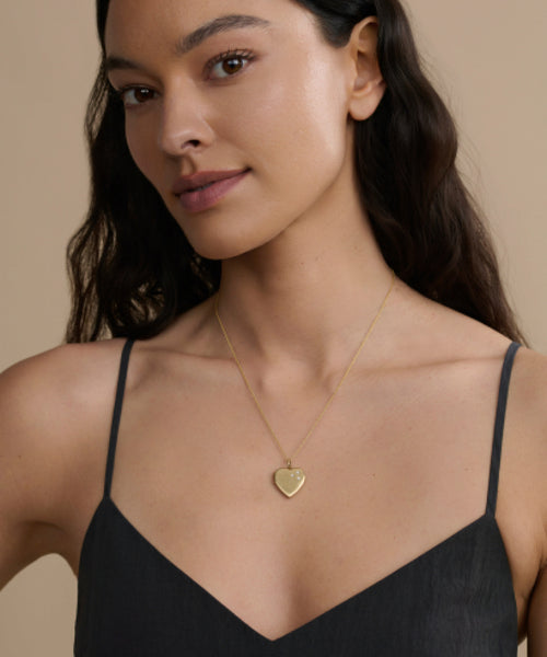 A woman with long dark hair wears a black spaghetti-strap top and the Bond Locket, posing against a beige background. The vintage-inspired Bond Locket adds timeless elegance to her look.