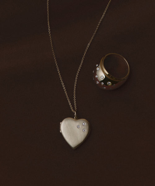 The Bond Locket, a gold heart-shaped necklace with delicate diamonds, rests beside a matching diamond-studded gold ring on a dark brown background—an elegant showcase of this vintage-inspired jewelry set.
