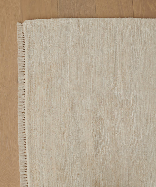 A close-up of the Bowery Rug, a beige handwoven piece with fringed edges, highlighting its subtle pattern and soft texture on a light wooden floor.