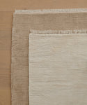 Two rectangular Bowery Rugs, one light beige and one brown, are layered on a light wood floor. The smaller beige Bowery Rug is placed diagonally over the larger brown Bowery Rug, both featuring fringed edges.