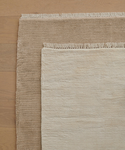 Two rectangular Bowery Rugs, one light beige and one brown, are layered on a light wood floor. The smaller beige Bowery Rug is placed diagonally over the larger brown Bowery Rug, both featuring fringed edges.