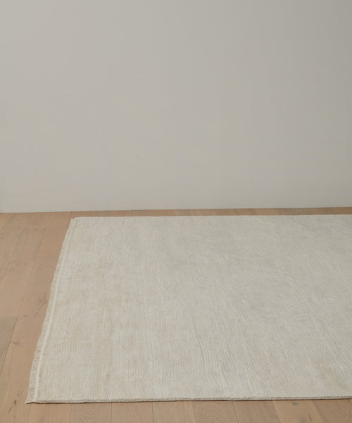 The Bowery Rug, a large handwoven rectangular piece in off-white, is displayed on a light wood floor against a plain white wall in a minimal, empty room.