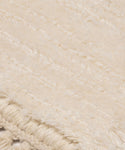 Close-up of the Bowery Rug, a cream-colored handwoven piece with a soft, fuzzy texture and subtle horizontal lines. The fringed edge in the bottom left corner features intricate braided details.