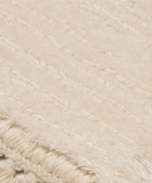 Close-up of the Bowery Rug, a cream-colored handwoven piece with a soft, fuzzy texture and subtle horizontal lines. The fringed edge in the bottom left corner features intricate braided details.