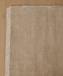 A close-up of the Bowery Rug in beige, handwoven with short fringe on the left edge. The rug displays subtle horizontal lines and a soft, plush look, shown atop a light wood floor.
