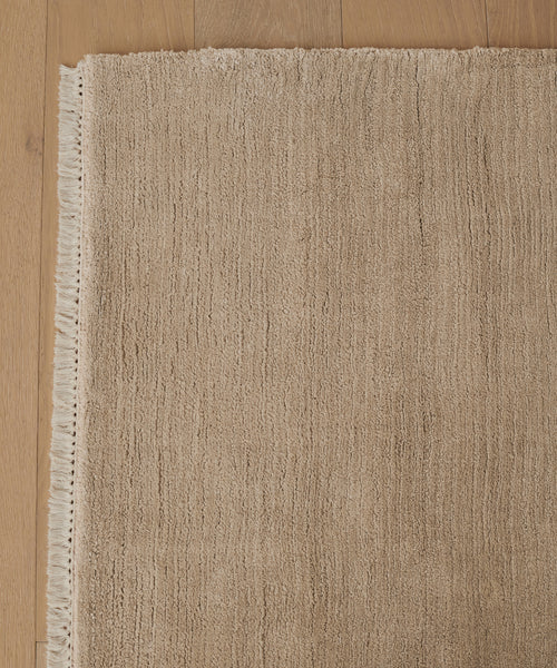 A close-up of the Bowery Rug in beige, handwoven with short fringe on the left edge. The rug displays subtle horizontal lines and a soft, plush look, shown atop a light wood floor.