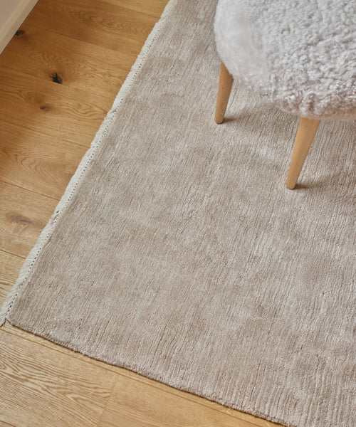 A close-up of the light beige Bowery Rug on a wooden floor, with the legs of a fuzzy white stool partially visible in the upper right corner.