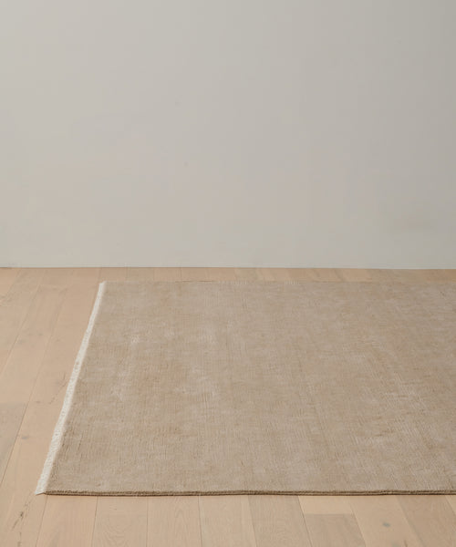 The Bowery Rug, a large rectangular handwoven beige rug, sits on a light wooden floor against an off-white wall in a minimalistic room.