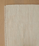 A close-up of a beige Rug Swatch with short fringes along the edge, displayed on a light wooden floor.
