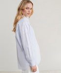 A woman wearing a light blue oversized long-sleeve button-up shirt, shown from a side-back angle against a plain light beige studio background.