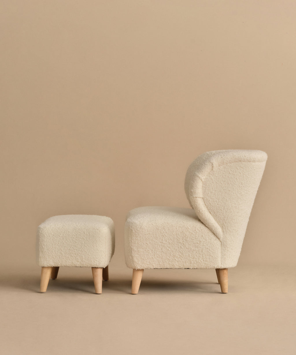 The Brentwood Chair in ivory wool boucle features a modern curved backrest and light wooden legs, paired with a matching ottoman, all set against a plain beige background.