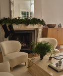 A cozy living room features a marble fireplace with greenery and two hanging stockings. Brentwood Chairs, a beige coffee table with a plant, and a wooden sideboard complete the elegant, neutral-toned space.