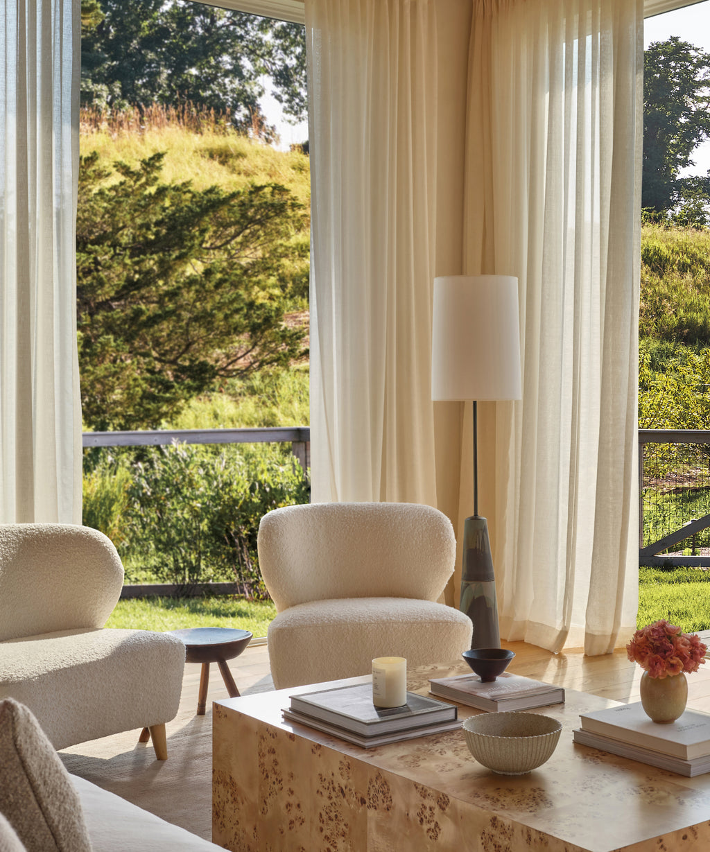 A bright, modern living room features cream Brentwood Chairs in Belgian Wool Boucle, a tall floor lamp, and a wooden coffee table with books, a candle, and flowers. Large windows showcase lush greenery outside.