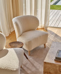 A cozy Brentwood Chair in ivory wool boucle sits beside a small wooden side table in a sunlit room with large windows, sheer curtains, a beige rug, and light wood floors. A coffee table and part of another chair are also visible.