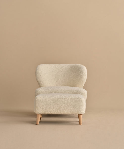 The Brentwood Ottoman, featuring light wood legs and cream-colored wool boucle upholstery, is centered against a plain beige background.