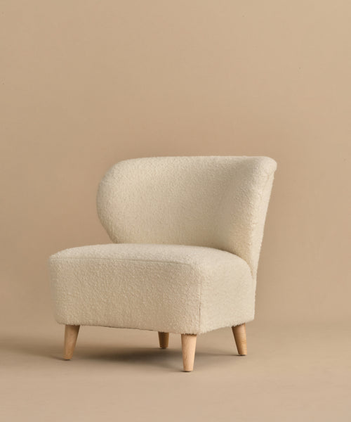 The Brentwood Chair features a modern armless design with a curved backrest, light wooden legs, and cream Belgian Wool Boucle upholstery, shown against a plain beige background.