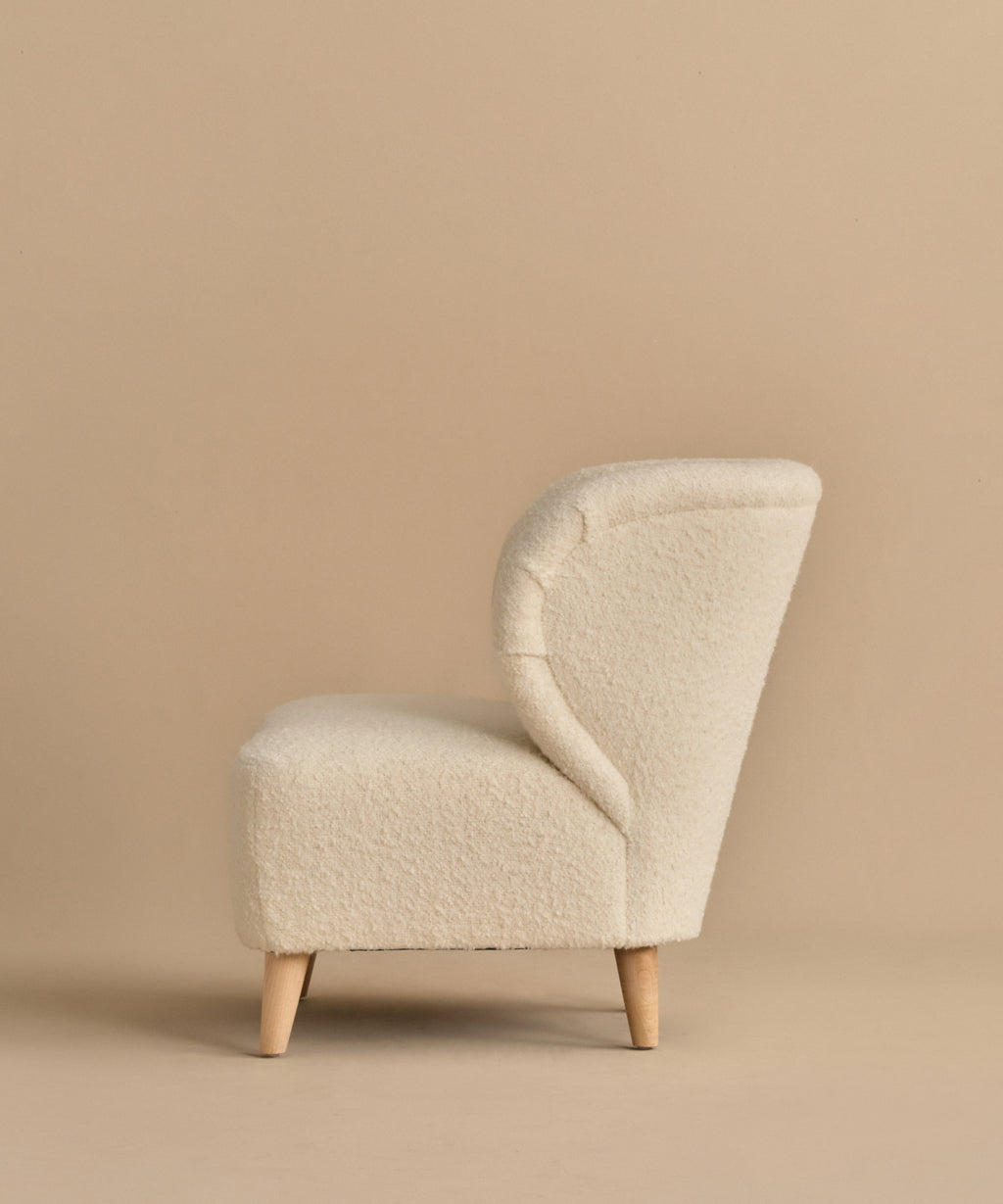 A side view of the Brentwood Chair, featuring ivory wool boucle upholstery and textured fabric with light wood legs, shown against a plain beige background.