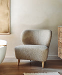 The Brentwood Chair, featuring textured Belgian wool upholstery and wooden legs, sits on a wooden floor beside a rustic dresser and white bathtub in a minimalist room with neutral-toned wall art partially visible on the left.