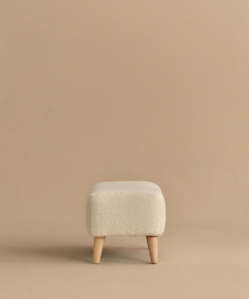 The Brentwood Ottoman is a small, square piece in cream wool boucle with textured fabric and four light wooden legs, shown on a plain beige background.