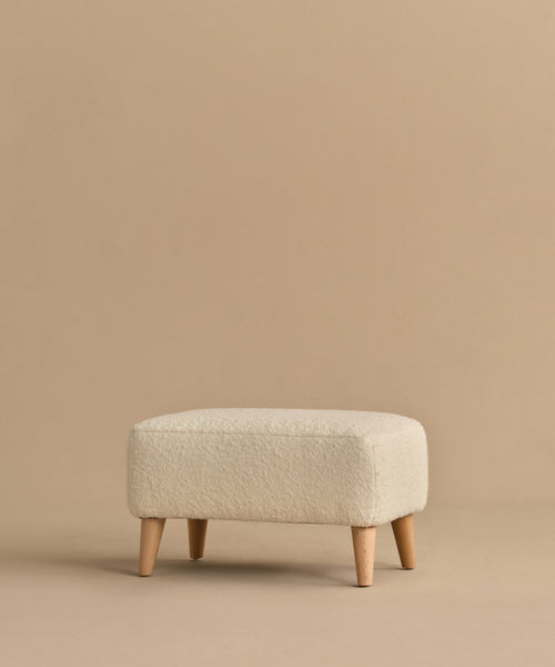 The Brentwood Ottoman features a minimalist design in light beige wool boucle with short wooden legs, showcased against a solid beige background.