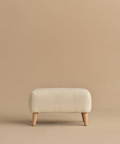 The Brentwood Ottoman is a compact, rectangular piece with off-white wool boucle upholstery and four short wooden legs, shown against a plain beige background.