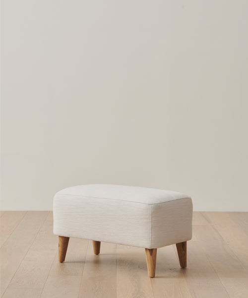 The Outdoor Brentwood Ottoman, small and rectangular with light beige upholstery and four wooden legs, sits on a light wood floor against a plain off-white wall.