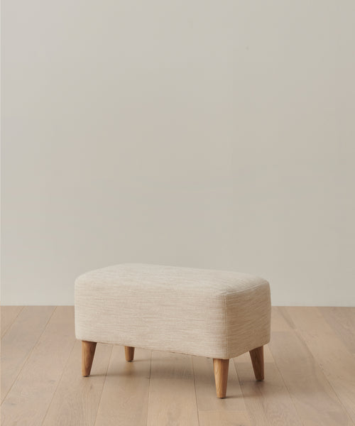 The Outdoor Brentwood Ottoman, featuring a small rectangular shape in light beige Performance Textured Linen and four solid teak legs, sits on a light wood floor against an off-white wall.